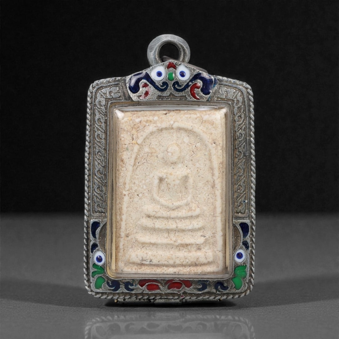 A Thai Pressed Powder Buddha Amulet in Enameled Metal Case, 20th Century, 45ZH: The amulet is formed from pressed powder, depicting a seated Buddha in bhumisparsha mudra, enclosed within a rectangular metal case with enameled decorative elements. The front of the case features bl