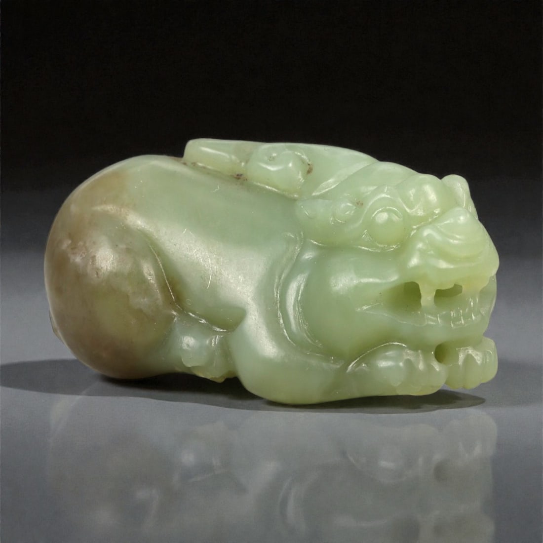 A Chinese Nephrite Jade Pixiu Carving, 20th Century, 95FA: Carved from pale celadon nephrite jade with areas of translucency and warm brown inclusions, the object is modeled in the round as a pixiu, the mythical guardian beast. The creature is shown recumbent