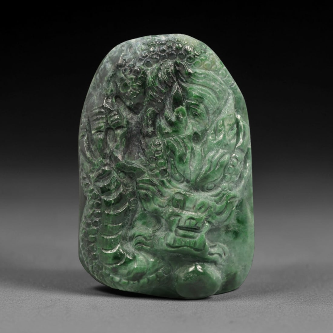A Chinese Jadeite Jade Pendant, 20th Century, 26ZL: Carved from green jadeite jade with natural dark green and greyish inclusions distributed throughout the matrix, the pendant is formed as an irregularly rectangular object with a relief-carved dragon