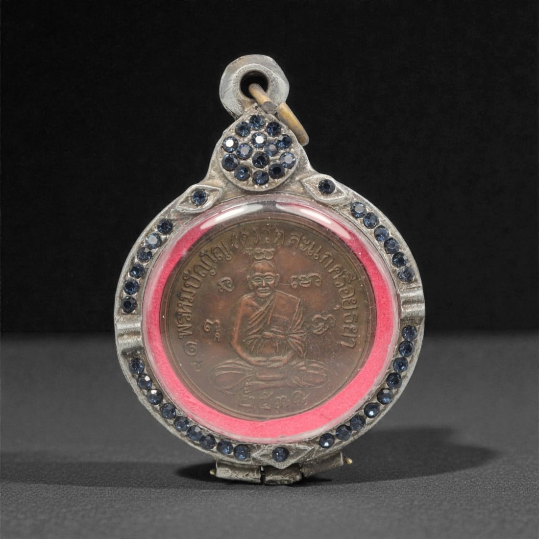 A Thai Monk Amulet Pendant with Blue Paste-Set Frame, 20th Century, 22FP: This circular amulet pendant features a patinated copper alloy core, encased in an ornate silver frame with a transparent protective cover, measuring 5.7 x 4 cm (2 1/4 x 1 9/16 in.). The obverse displ