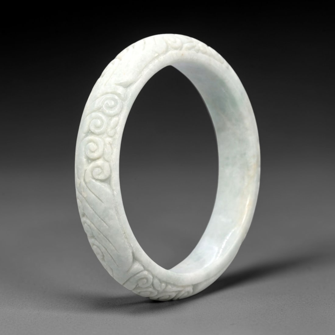 A Chinese Jadeite Jade Bangle Bracelet, 20th Century, 50FS: Carved from pale celadon jadeite jade with a smooth, polished finish, the bangle is formed as a continuous circular band. The exterior surface is decorated with raised relief carving depicting scrolli