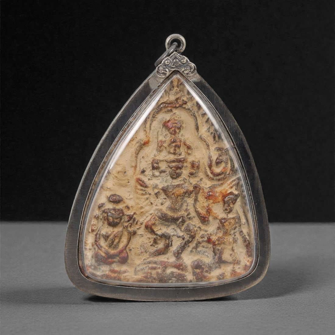 A Thai Multi-Armed Deity Amulet with Silvered Frame, 20th Century, 39FP: The amulet is of triangular form, featuring a molded multi-armed deity figure on the obverse, rendered in a dark patinated material against a light beige ground. The deity is depicted in a dynamic pos