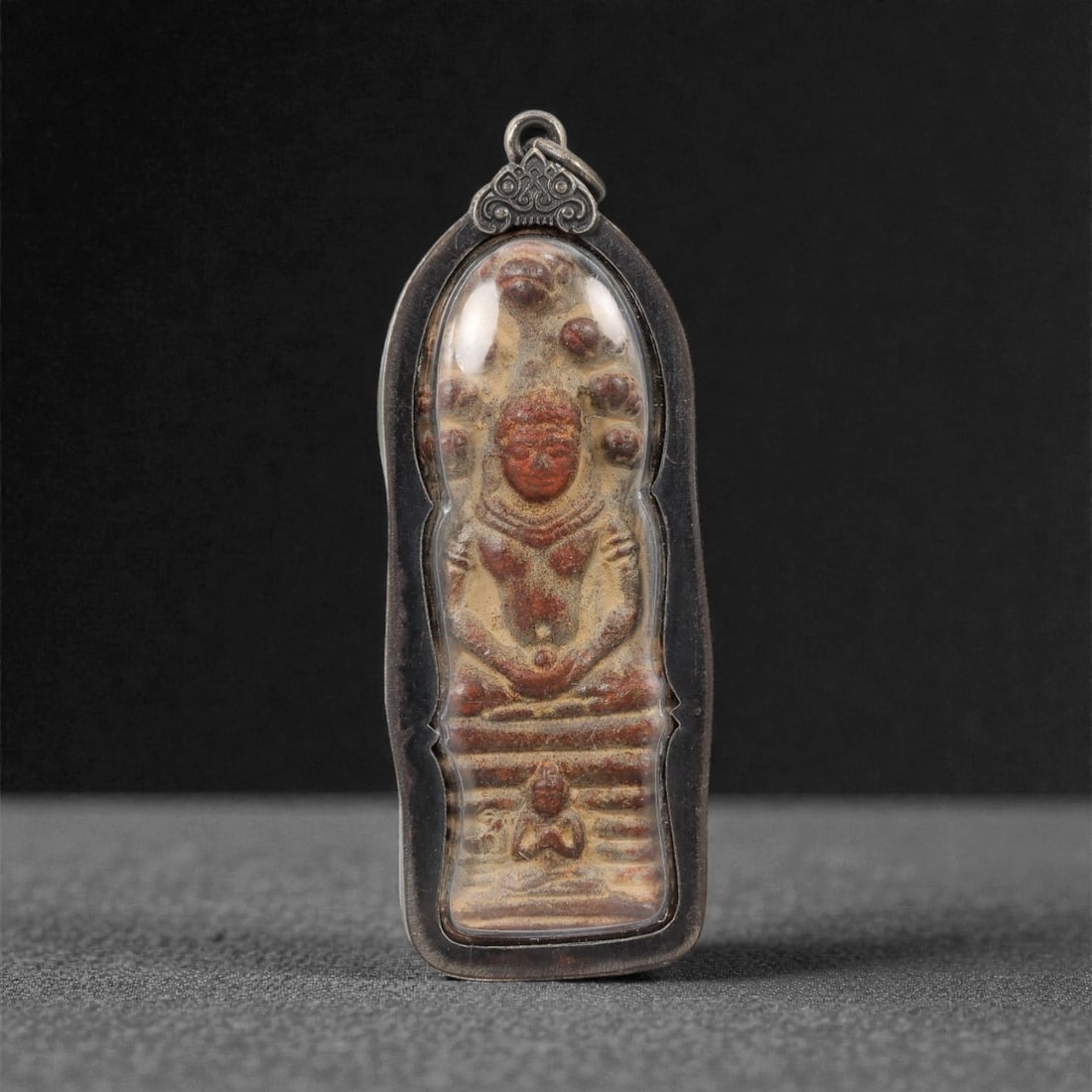 A Thai Seated Deity Amulet with Metal Frame, 20th Century, 29FP: The amulet is of elongated, teardrop-shaped form, featuring a molded seated deity figure on the obverse, rendered in a dark patinated material against a light beige ground. The deity is depicted with