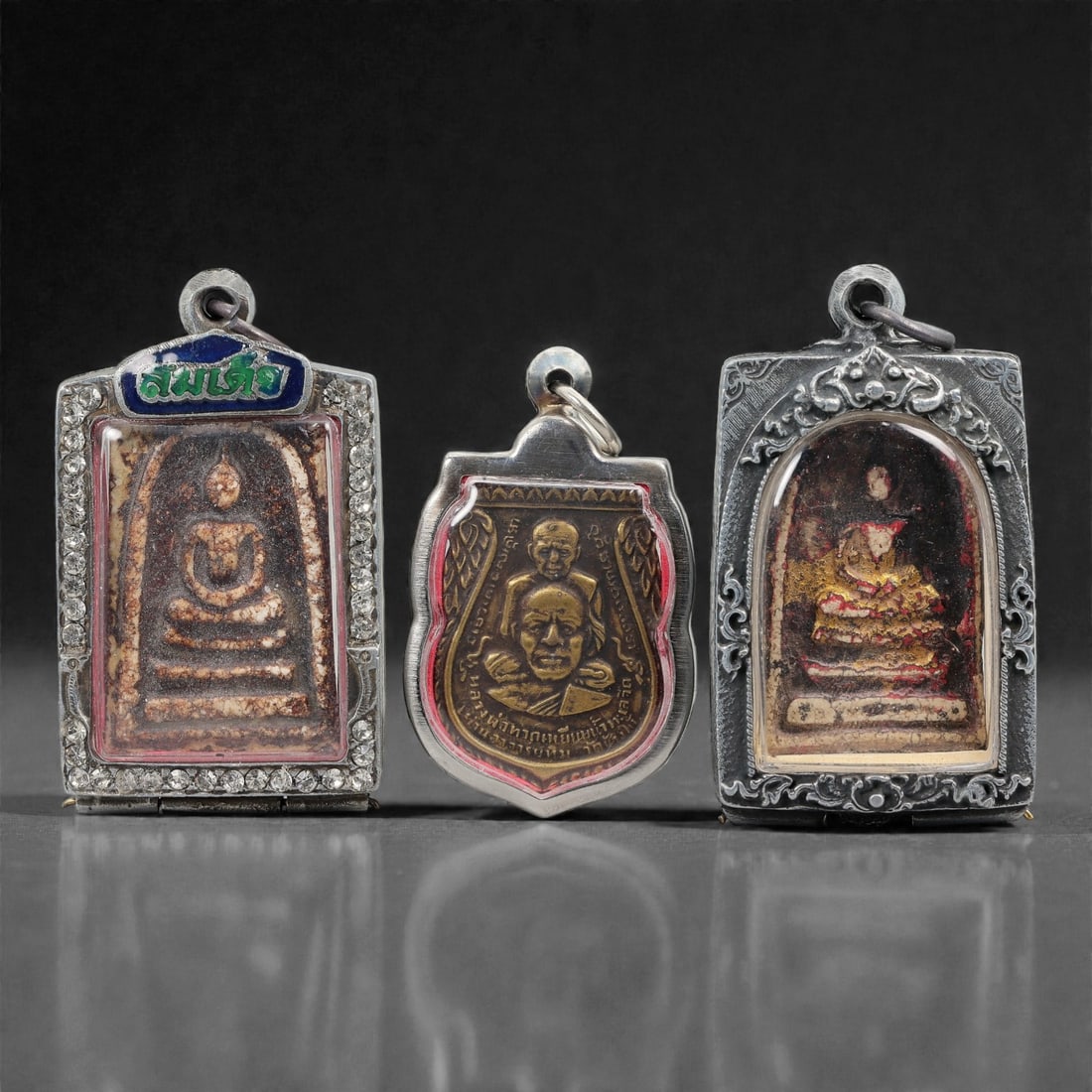 A Group of Three Thai Vintage Buddhist Amulets, 20th Century, 91FM: Comprising three Thai Buddhist amulets, each encased in ornate metal frames with visible surface patina and wear. The leftmost amulet is a rectangular phra somdej-style tablet, featuring a seated Budd