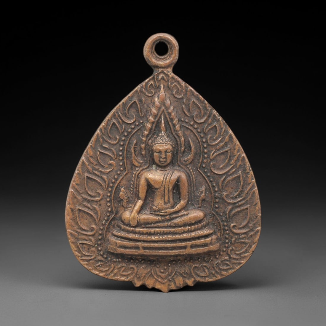 A Thai Copper Alloy Buddha Amulet, Late 20th Century, 93FP: A teardrop-shaped copper alloy amulet, measuring 3.5 x 2.8 cm. The obverse depicts the Buddha seated in meditative posture on a multi-tiered pedestal, framed by ornate scrollwork and flame motifs. The