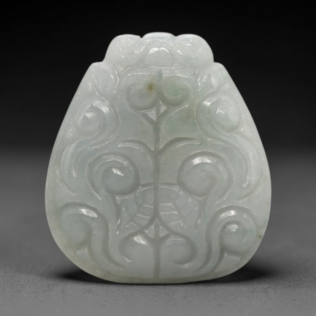 A Chinese Jadeite Jade Scrollwork Pendant, 20th Century, 89FR: Carved from translucent white jadeite jade with faint pale green internal veining, the pendant features a symmetrical scrollwork pattern in low relief. A central vertical incised line divides the comp