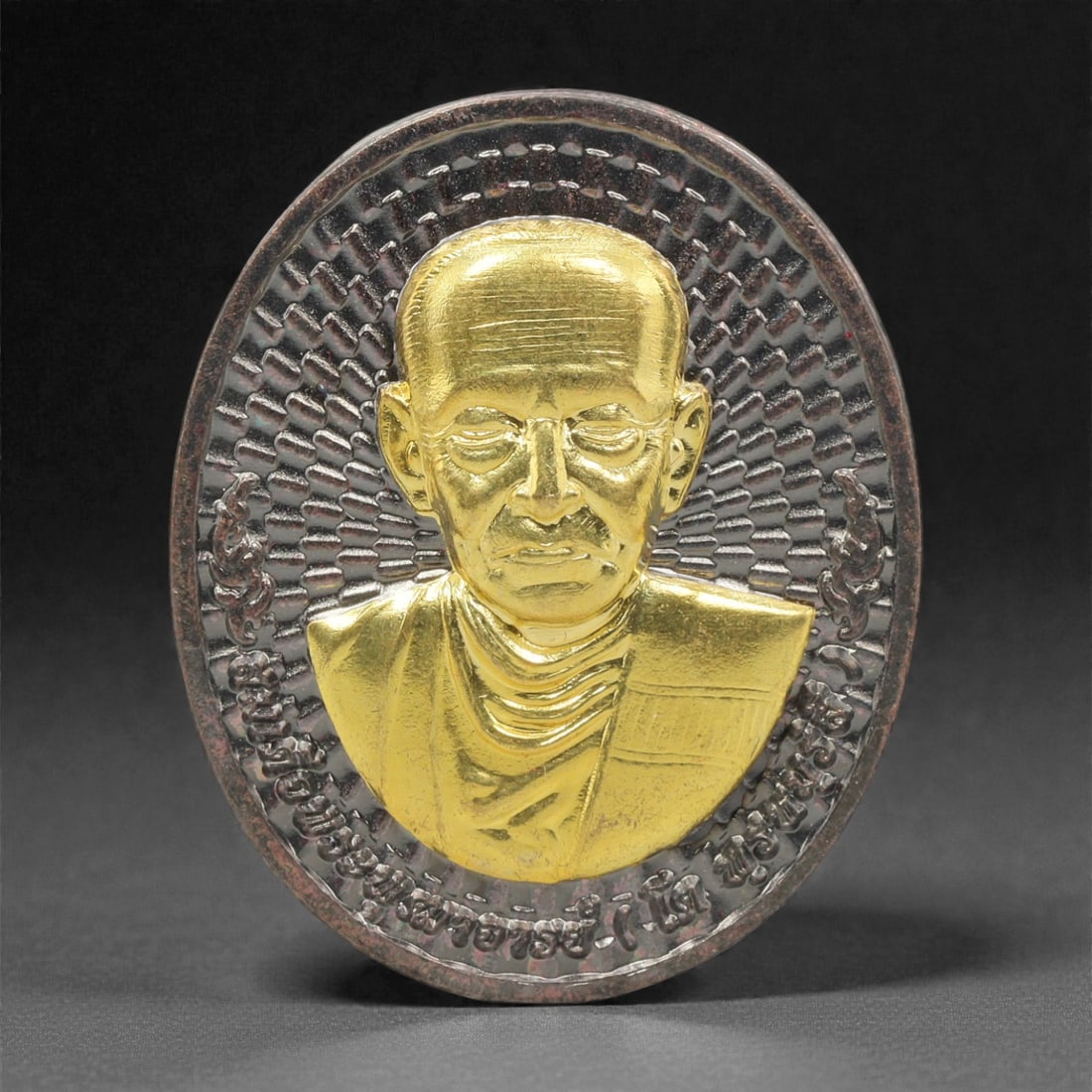 A Thai Bi-Metallic Monk Portrait Medallion, 21st Century, 86FP: Cast with a bi-metallic construction, the medallion is oval and double-sided with a raised dark metal border. The obverse features a high-relief gilt bust portrait of a Buddhist monk, set against a da