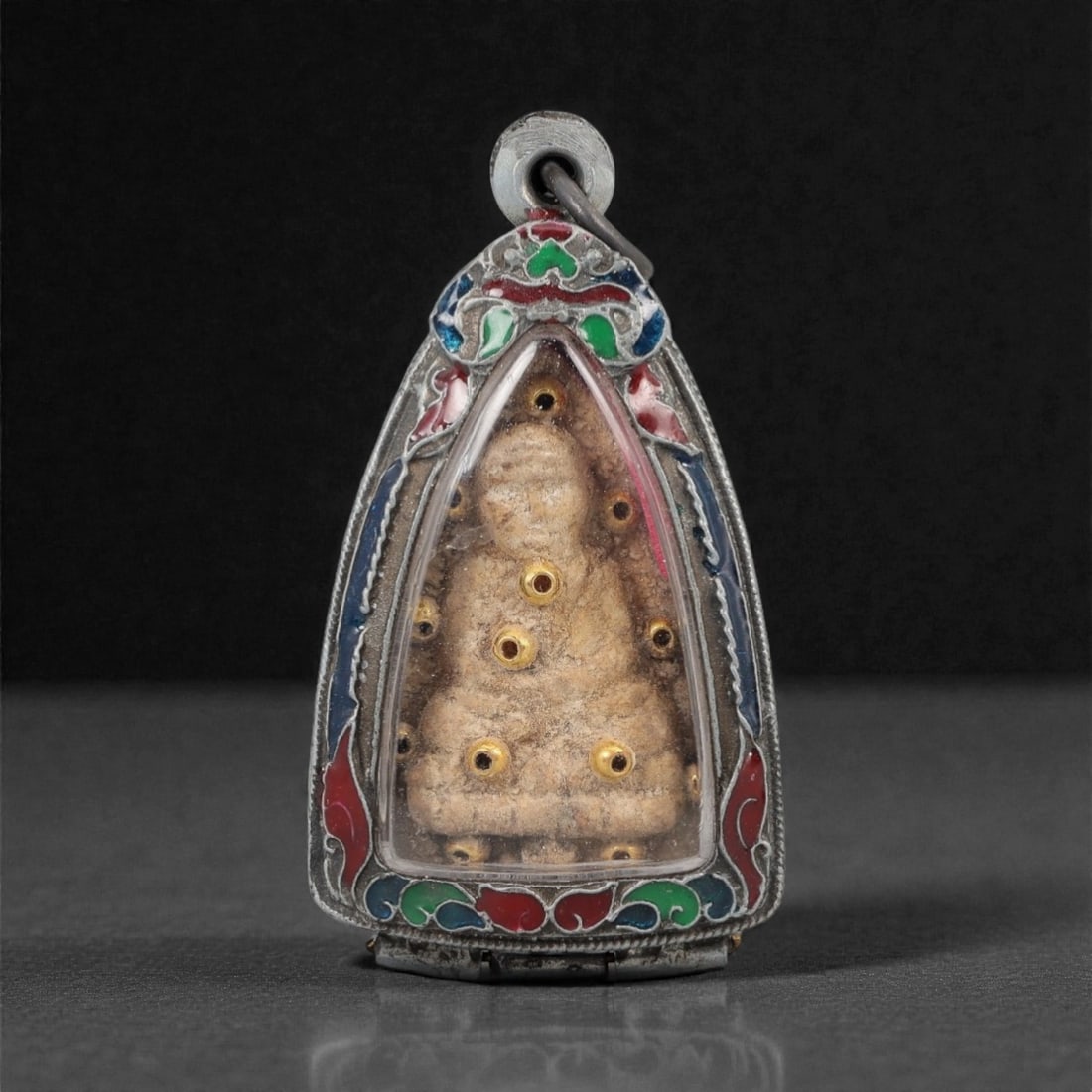 A Thai Seated Monk Amulet with Enameled Metal Frame, 20th Century, 30FP: The amulet is of triangular form, featuring a molded seated monk figure on the obverse, rendered in a light beige composite material with embedded gold-colored metal inlays. The reverse displays a pla