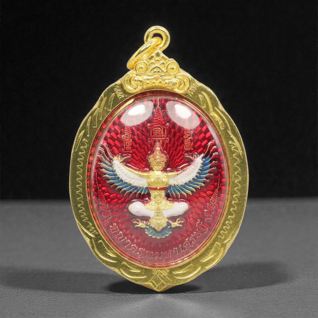 A Thai Gilt-Metal Garuda and Rahu Amulet, 21st Century, 78FP: Constructed with a cast gilt-metal core encased in a matching gilt-metal bezel fitted with an integral suspension loop at the upper apex, the amulet features a double-sided design secured beneath clea