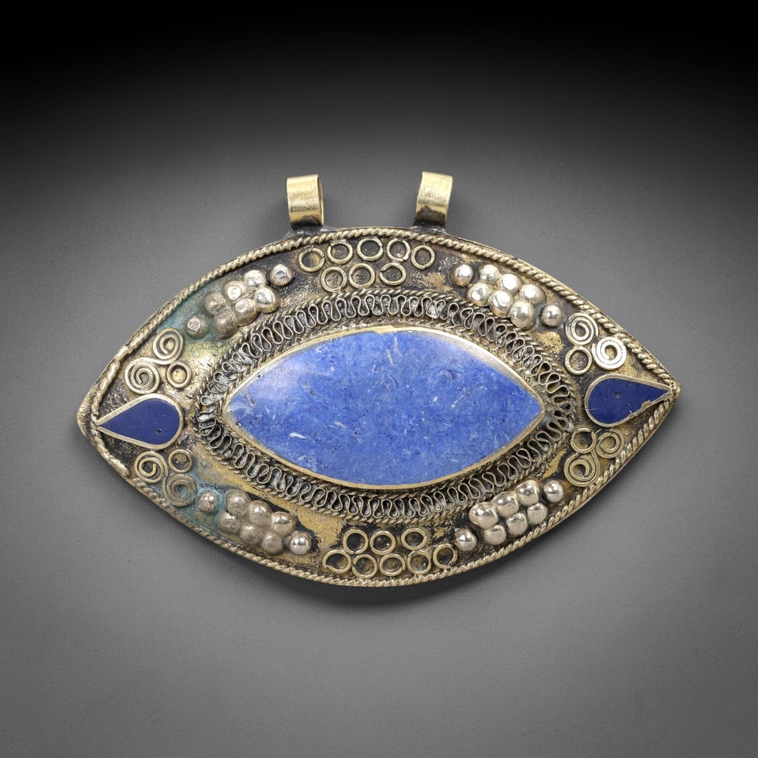 A Tibetan Brass and Silver Pendant with Lapis Lazuli Inlay, 20th Century, 94XM: Crafted from brass with silver repousse and wirework, the pendant has an eye-shaped form with two cylindrical suspension loops at the top. The central element is a large, polished lapis lazuli cabocho