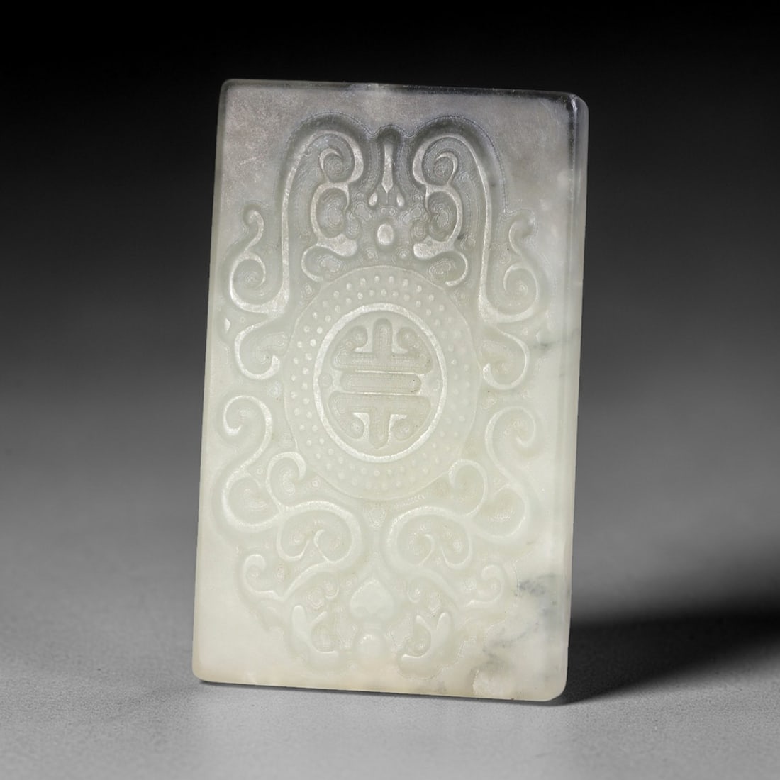A Chinese Hetian Jade Plaque Amulet, 20th Century, 79FH: Carved from pale celadon Hetian jade with natural greyish inclusions and subtle veining, the plaque is rectangular with bevelled edges. The surface is rendered in low relief, featuring a symmetrical c