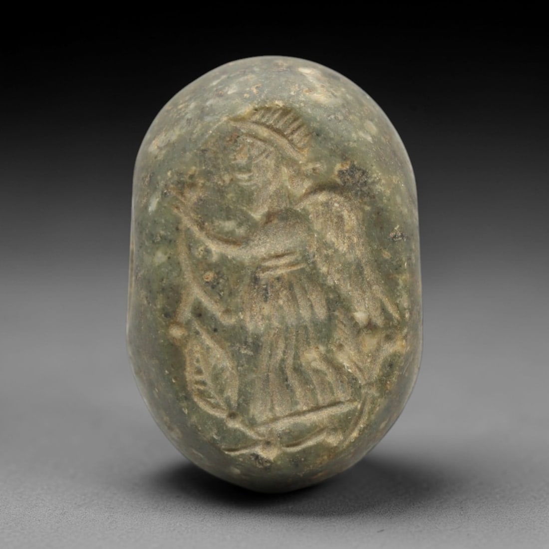 Ancient Stone Hemispherical Stamp Seal - Near Eastern Winged Figure Amulet, 14ZS: This ancient Near Eastern hemispherical stamp seal is carved from a muted green stone, likely serpentine or steatite, with a weathered, earthy patina and subtle mineral inclusions. The seal's oval fac