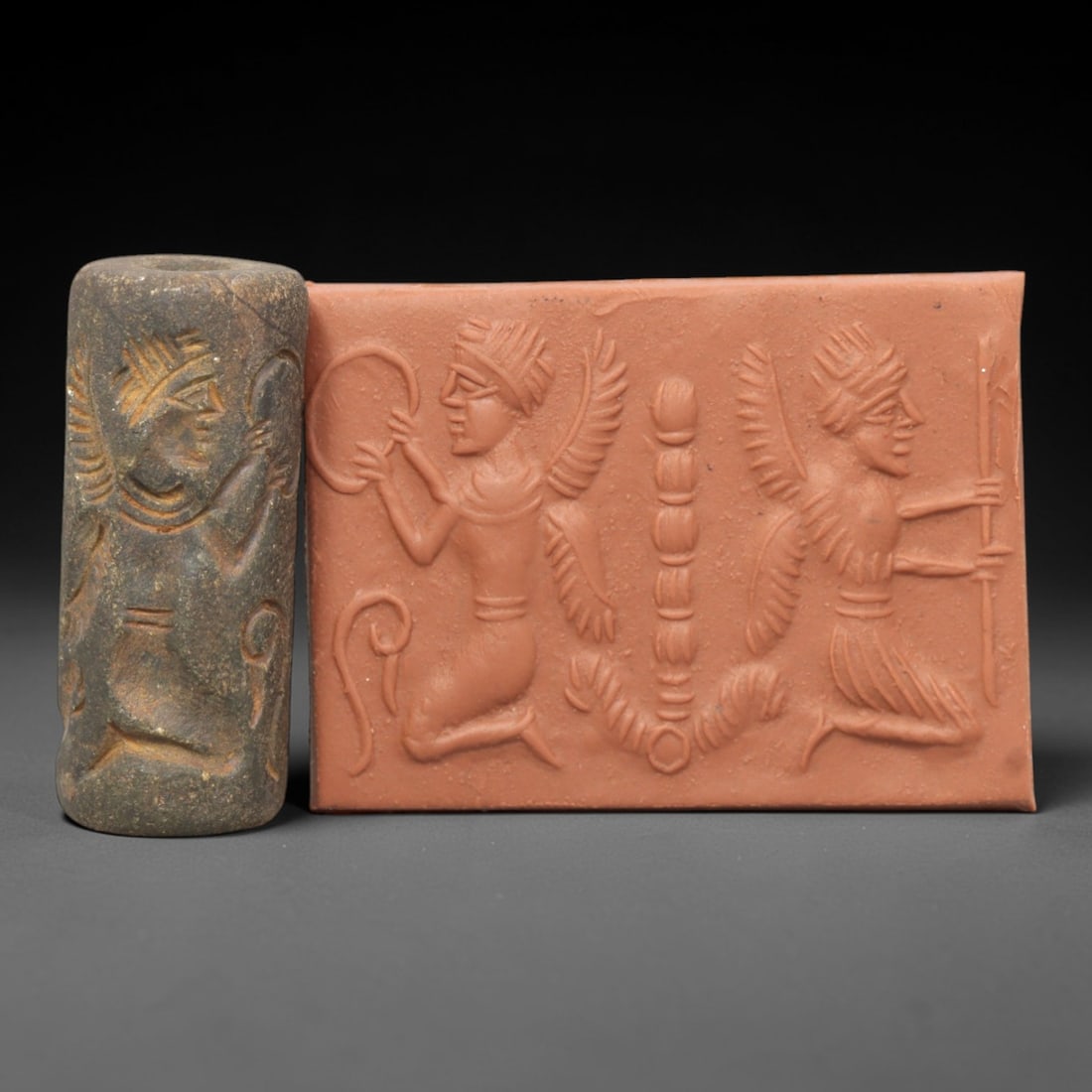 Ancient Steatite Cylinder Seal - Mesopotamian Winged Attendants Ritual Scene, 60AL: Carved from dark, granular steatite, this ancient Mesopotamian cylinder seal depicts a symmetrical ritual scene, executed with precise glyptic craftsmanship characteristic of the Late Bronze Age. The
