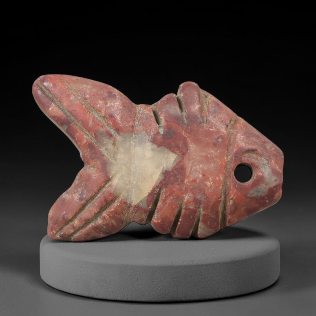 Ancient Stone Fish Amulet - Egyptian Ritual Figurine, 70AG: This amulet is carved from a single piece of mottled red stone, exhibiting a granular texture with a prominent pale mineral inclusion at its center and subtle ochre patina across its surface. The form