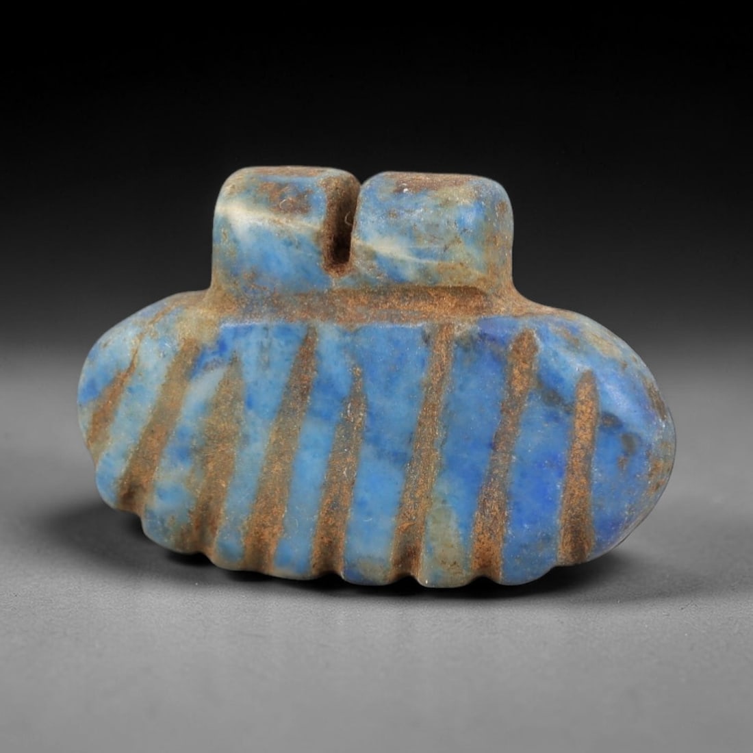 Ancient Lapis Lazuli Amulet - Egyptian Winged Pendant, 26AP: Carved from lapis lazuli, this amulet features a rounded, wing-like form with two raised upper lugs framing a central suspension channel, its surface incised with vertical bands that define its segmen