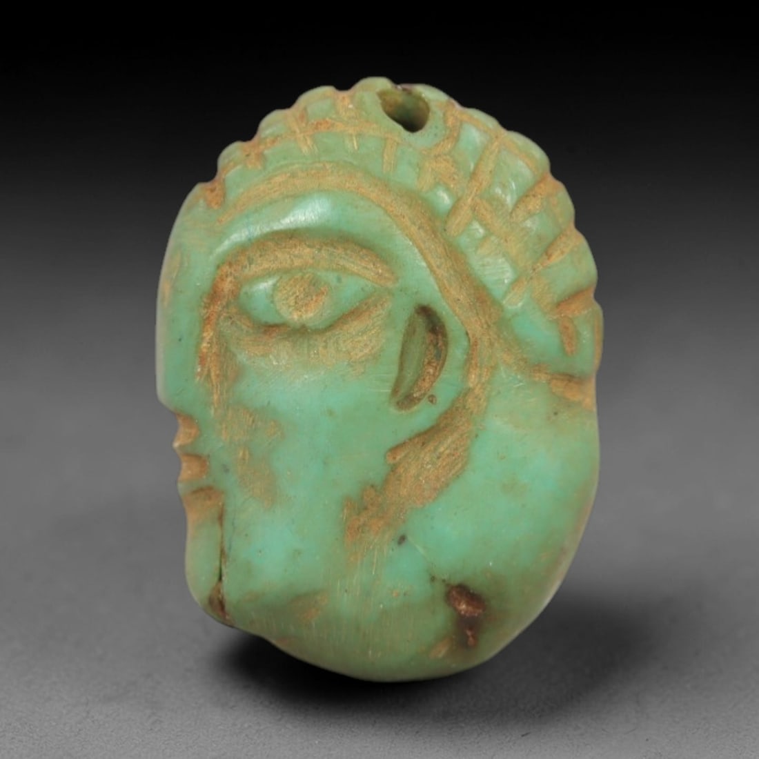 Ancient Turquoise Face - Near Eastern Amulet, 87AK: Carved from turquoise, this amulet features a stylized human face with incised detailing defining the eye, ear, and textured hair or headdress, with a suspension hole at the top for wear. Attributed t