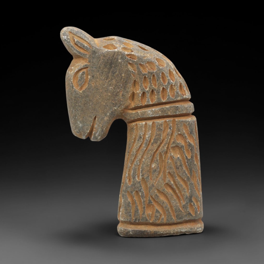 Ancient Near Eastern Stone Horse Head Amulet - Figurine, 37ZR: This artifact is a carved stone horse head amulet, crafted from a granular, grey-brown stone with a textured surface and earthy patina filling the incised decorative details. The piece depicts a styli