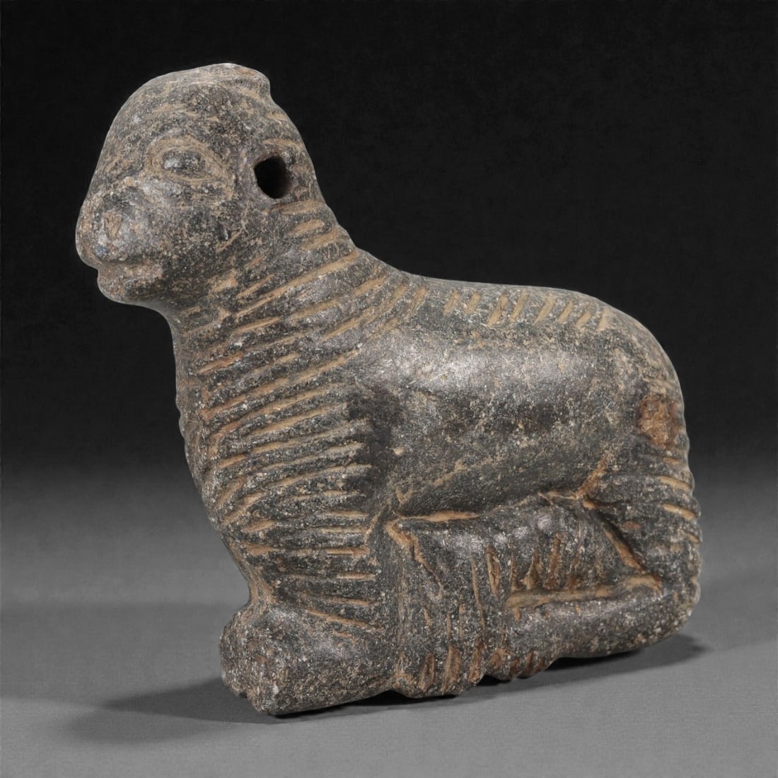 A Mesopotamian Schist Recumbent Ram, Early 2nd Millennium BCE, 61LK: Carved from dark grey schist, the figure depicts a ram in a recumbent posture with the legs tucked beneath the body and the head raised forward. Incised parallel lines articulate the fleece along the