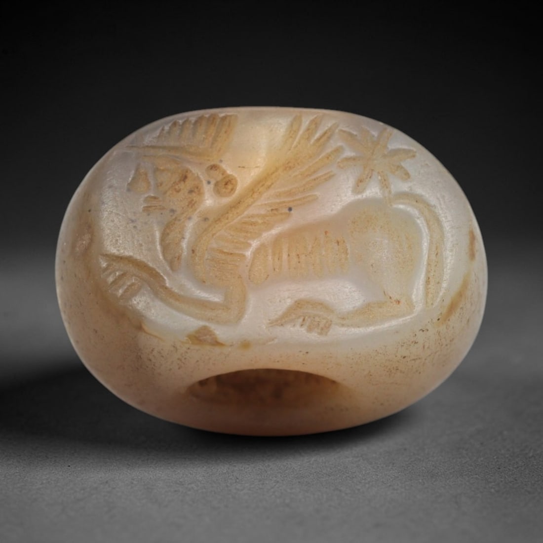 Ancient Chalcedony Stamp Seal - Near Eastern Winged Sphinx, 41ZS: Crafted from translucent chalcedony, this hemispherical stamp seal bears an incised scene of a winged sphinx-like figure in profile, accompanied by a stylized palm tree motif, executed with precise li