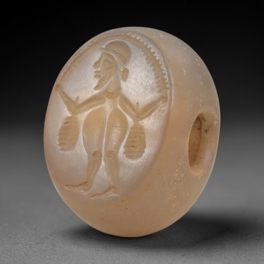 Ancient Chalcedony Stamp Seal - Sasanian Figural Bearded Male, 67AK: Carved from translucent chalcedony, this hemispherical Sasanian stamp seal features a central standing bearded male figure, holding two hanging bundles in outstretched hands, beneath a radial decorati