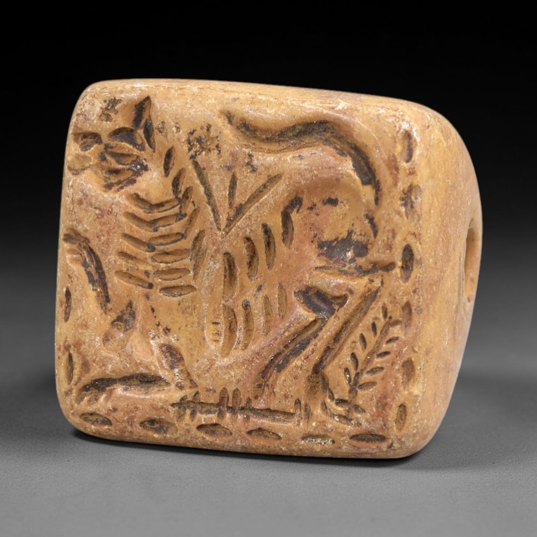 Ancient Stone Stamp Seal - Near Eastern Animal Motif, 72AL: Carved from tan limestone, this triangular stamp seal features a raised impression face depicting a dynamic animal scene, likely a hunting or ritual interaction, with incised linear motifs defining th