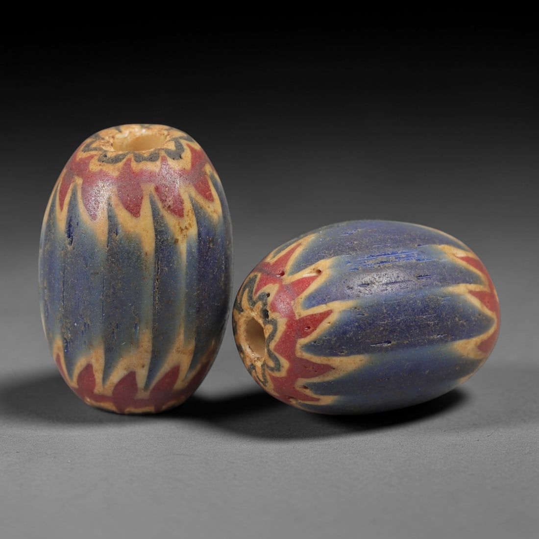 A Pair of Venetian Glass Chevron Beads, 18th-19th Century, 82AC: Crafted from translucent glass with a pale yellow core, each bead is decorated with a chevron pattern of alternating deep blue and vibrant red layers, radiating from the central perforation to the out