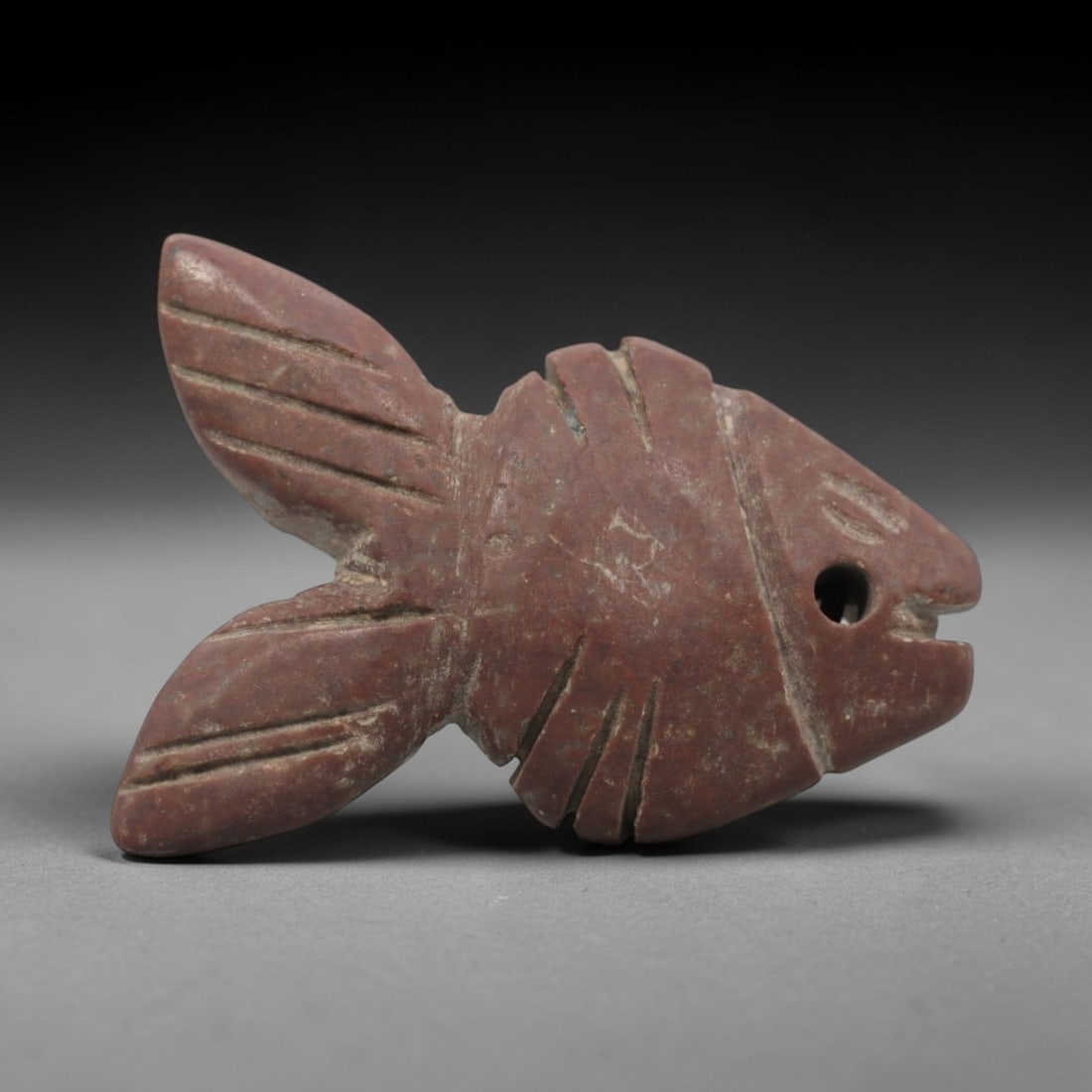 Ancient Egyptian Stone Fish Amulet - Carved Pendant Figure, 15ZV: This amulet is carved from a dense, reddish-brown stone, likely siltstone or a similar sedimentary rock, with incised linear details defining its form. The piece depicts a stylized fish, with a dorsal