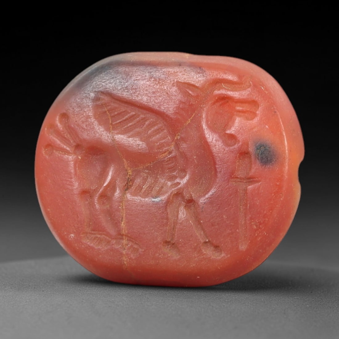 Ancient Carnelian Intaglio - Near Eastern Winged Beast Seal, 16AL: Crafted from rich red carnelian, this circular intaglio bears a deeply incised depiction of a winged quadrupedal beast with horned features, accompanied by a vertical sword-like symbol to its right. T