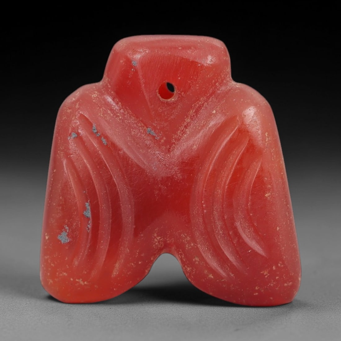 Ancient Carnelian Bird Amulet - Egyptian Figurine, 29AJ: Carved from vibrant red carnelian, this amulet is formed as a stylized bird with outstretched wings, featuring incised curved linear detailing across each wing to denote feather patterns. A central su