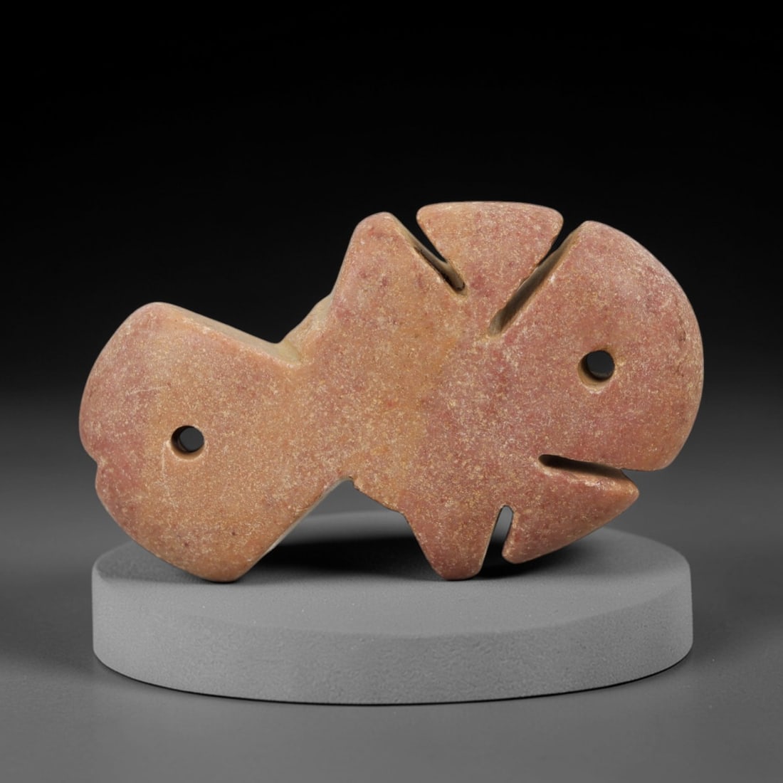 Ancient Stone Fish Amulet - Egyptian Carved Pendant, 45AE: This amulet is carved from a light reddish-tan stone with a fine, granular texture and naturally weathered patina. The form is a stylized fish, with angular incised notches defining the dorsal, ventra