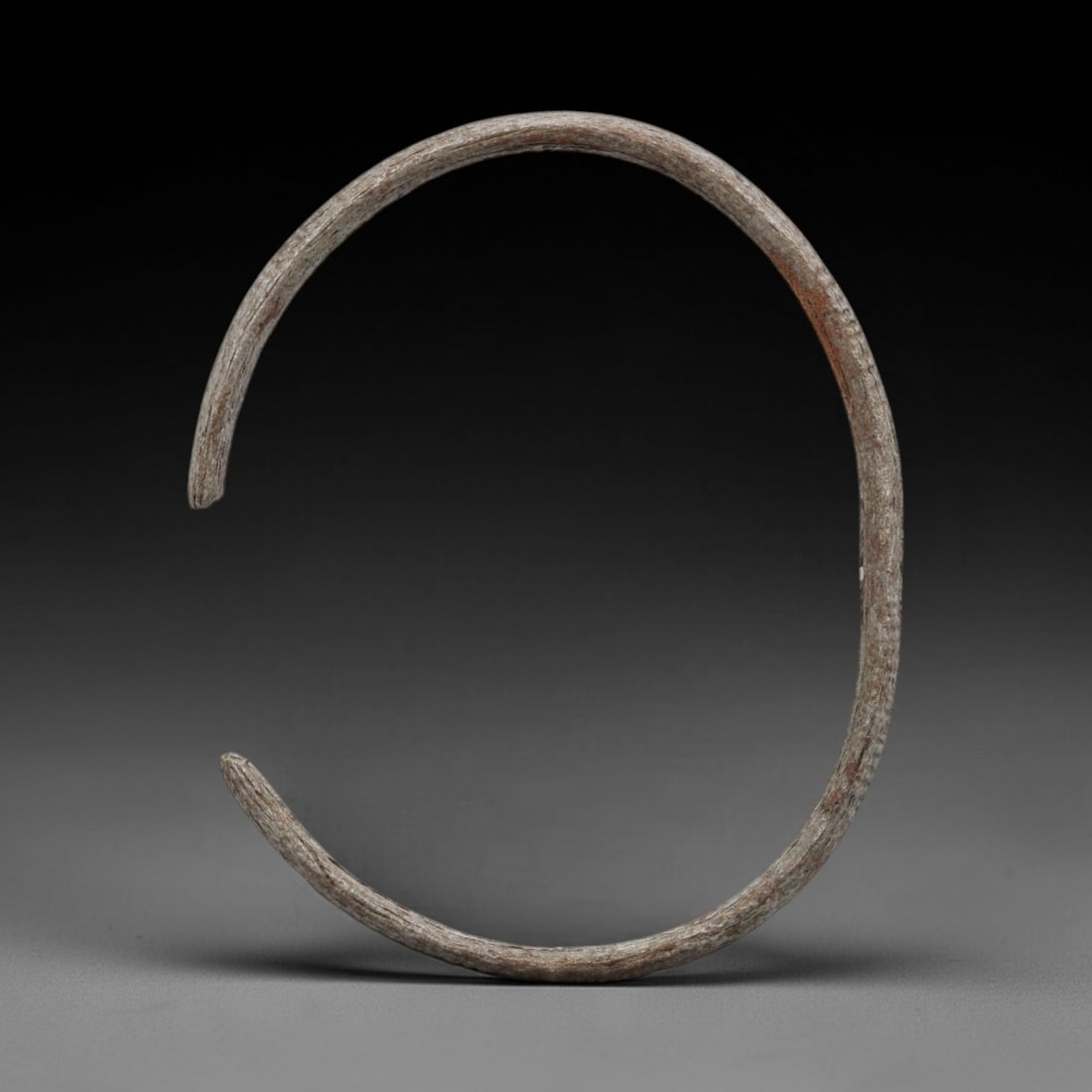 A Celtic Iron Penannular Bracelet, 2nd Century BCE - 1st Century CE, 3FV: Forged from iron with a fine, consistent rounded section, the bracelet is shaped into a pronounced open penannular form with a broad terminal gap and simple, unbevelled ends. The surface presents a te