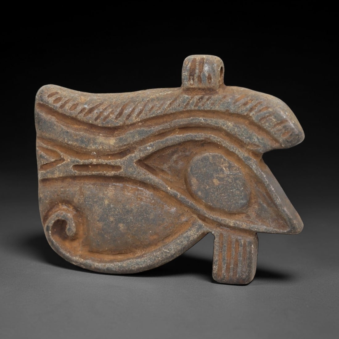 Ancient Stone Wedjad Eye - Egyptian Amulet Figurine, 19AM: This Wedjad Eye amulet is carved from a dense, dark stone with a mottled, earthy patina showing subtle ochre discoloration and fine surface striations. The form depicts the iconic Eye of Horus, featur
