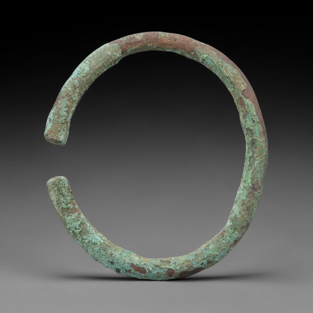 A Roman Bronze Bracelet, 1st-3rd Century CE, 6FV: Cast from bronze with a thick, rounded section, the bracelet is formed as an open C-shape with rounded, slightly tapered terminals. The surface is covered in a patchy green patina with extensive reddi