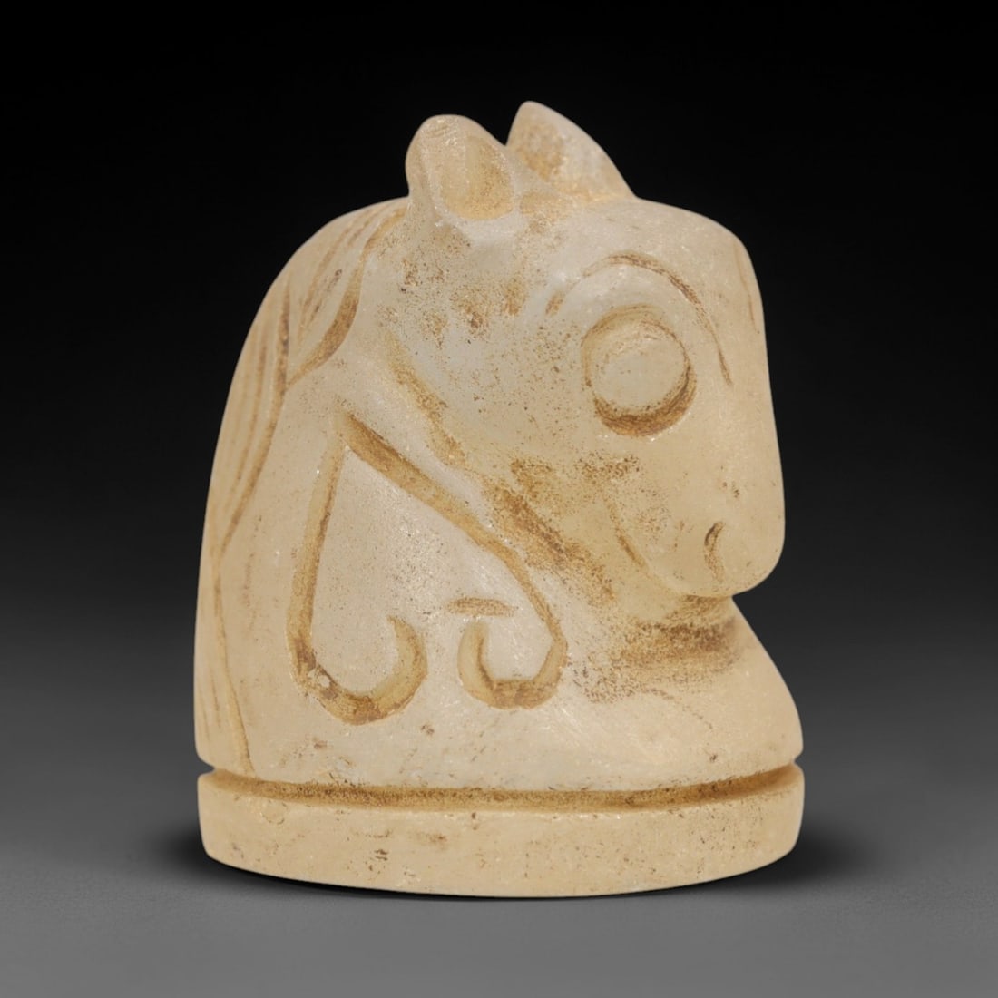 Ancient Medieval Egyptian Glass Horse Chess Piece, 74ZR: Carved from translucent pale glass with earthy encrustations, this chess piece depicts the head and neck of a horse, featuring stylized linear details defining the ears, eye, mane, and decorative harn