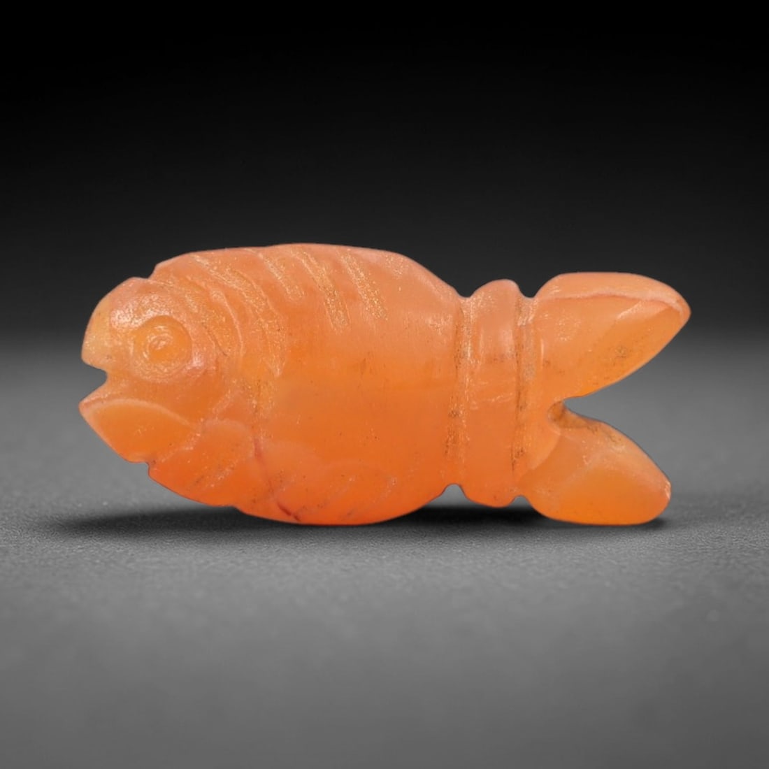 Ancient Carnelian Fish Amulet - Egyptian Figurine, 48XS: Carved from warm, translucent orange-red carnelian with natural tonal gradients and subtle surface patination, this amulet is formed as a stylized fish with incised linear detailing to define segmente