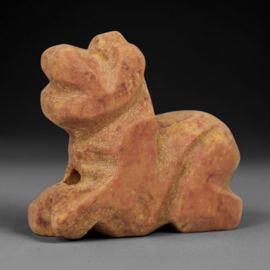 Ancient Reddish Stone Recumbent Feline - Near Eastern Amulet, 12AE: Carved from warm reddish-tan stone with natural granular texture, this amulet depicts a recumbent feline in a compact, stylized form. The creature is rendered in a prone resting posture, with simplifi