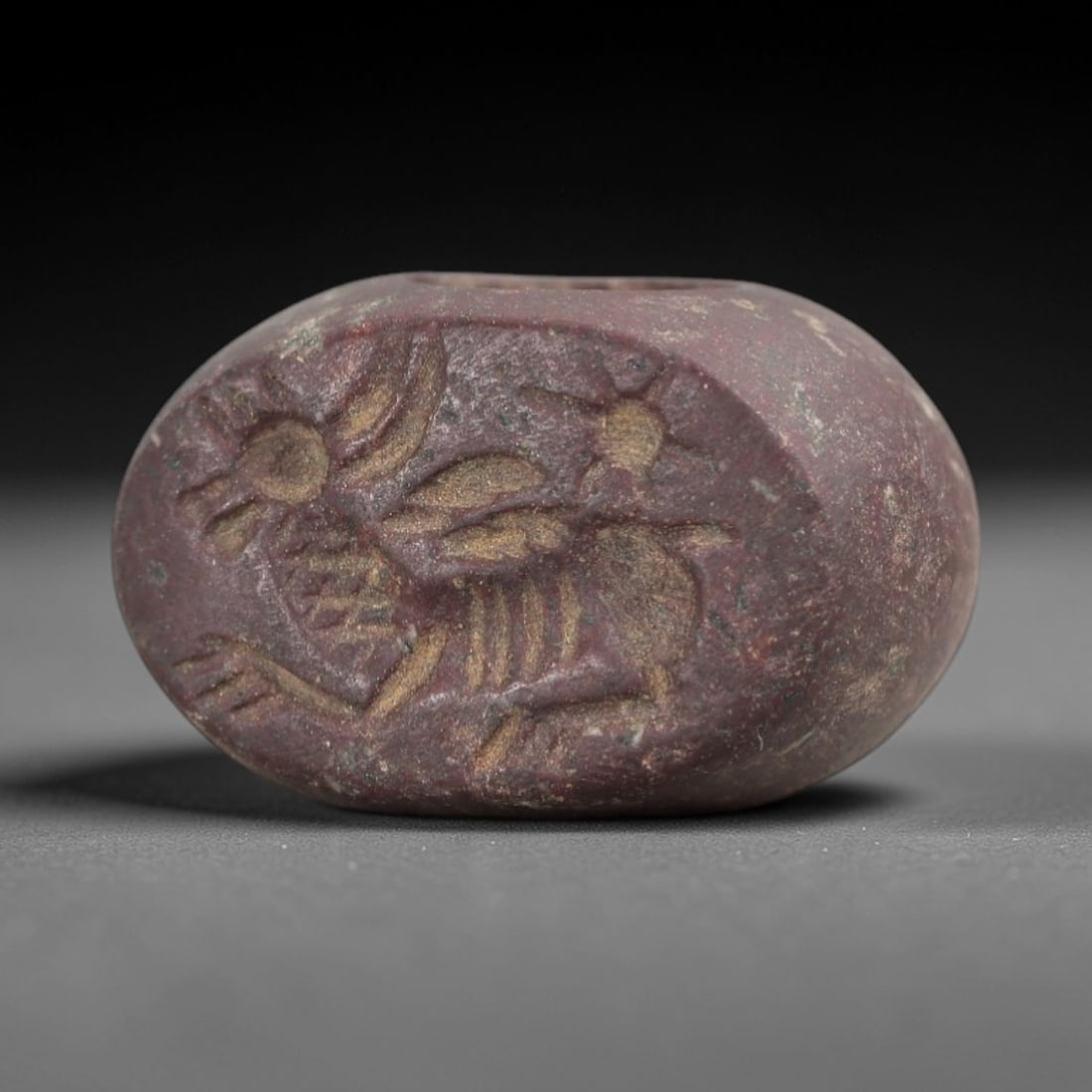 Ancient Stone Hemispherical Stamp Seal - Near Eastern Winged Stag Figure, 21AN: Carved from a dense, dark reddish-brown stone, this hemispherical stamp seal features a flat, oval face incised with a stylized winged stag in profile, accompanied by a radiate sun motif. The stag is