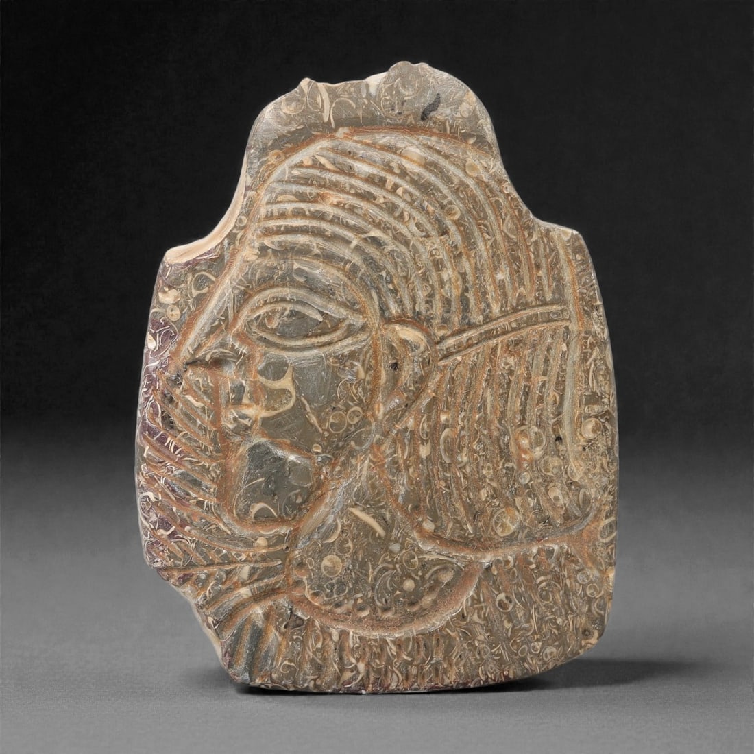 A Sasanian Fossilized Stone Plaque, 3rd-7th Century CE, 78FL: Carved from fossilized stone with natural inclusions and reddish-brown incised decoration, the object is formed as an irregularly shaped plaque with a relief portrait of a male figure in profile. The