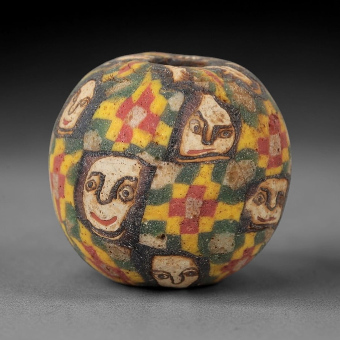 Ancient Glass Face Bead - Egyptian Amulet Bead, 50ZM: This ancient bead is crafted from polychrome glass, formed using core-formed and trailed decorative techniques characteristic of Egyptian glassworking traditions. The spherical form features multiple