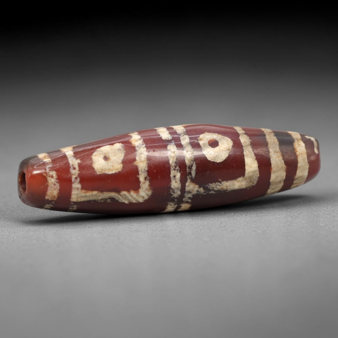 Ancient Carnelian 2-Eye Dzi Bead - Himalayan Amulet, 65XV: Crafted from rich translucent carnelian, this bead features two etched circular eye motifs paired with linear and geometric bands, executed in the traditional Himalayan Dzi bead technique. The form is