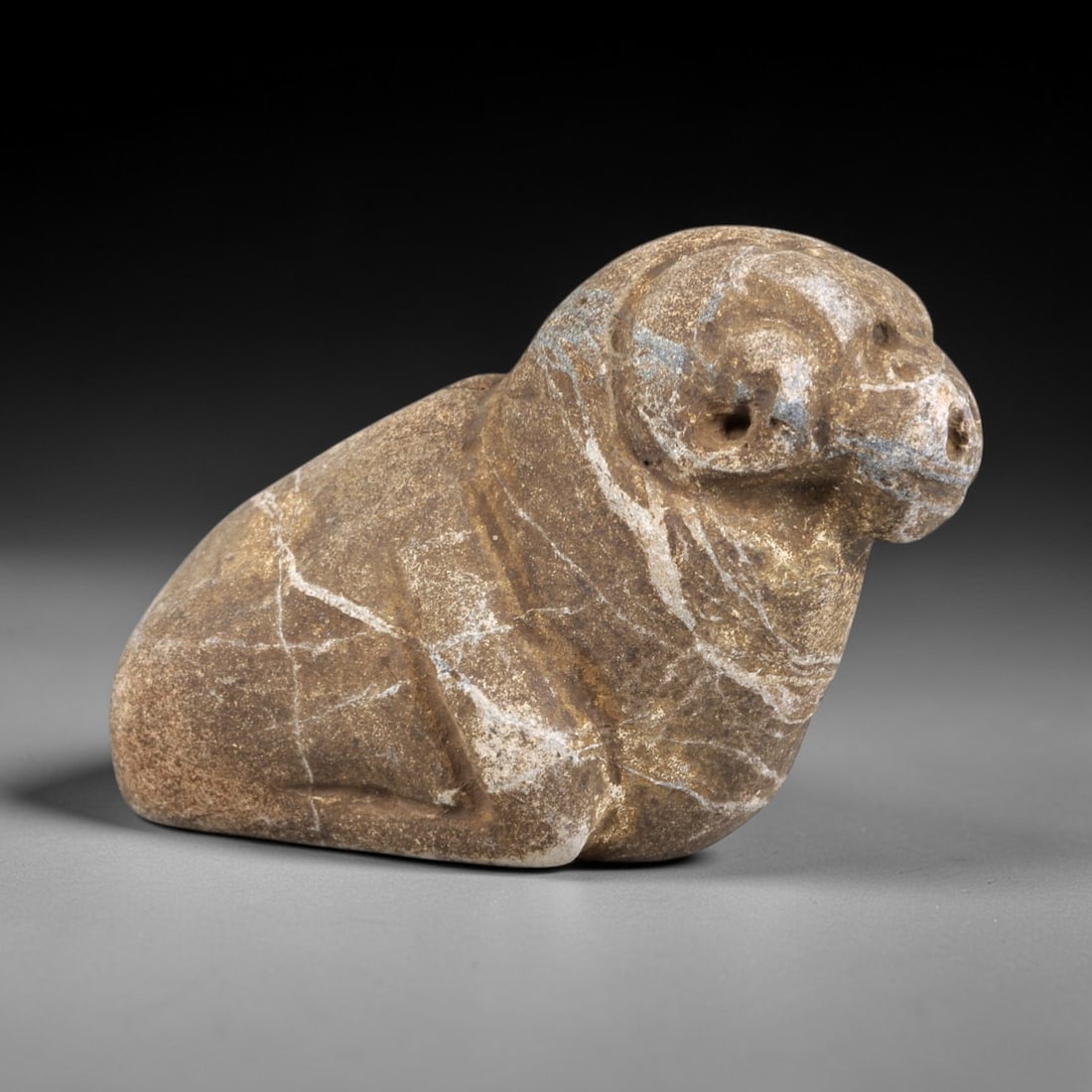 Ancient Stone Ram Effigy - Near Eastern Zoomorphic Figurine, 88FX: Carved from a veined, granular stone, this effigy depicts a recumbent ram in a compact, stylized form. Relief details define the curled horns, snout, and rounded body, with small perforations in the f