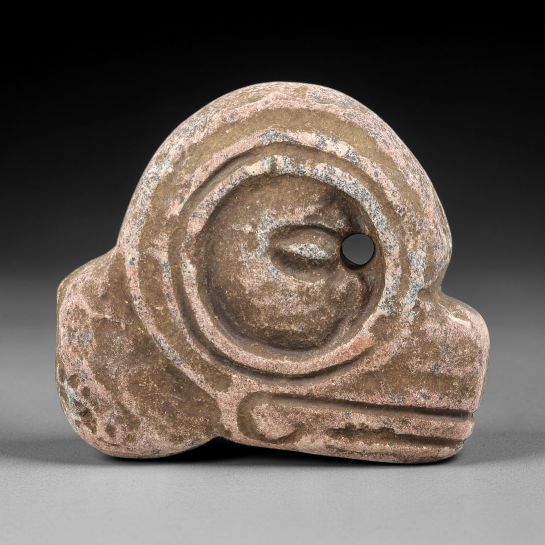 Ancient Stone Anthropomorphic Amulet - Near Eastern Figurine, 96FX: Carved from a speckled, granular stone, this anthropomorphic amulet features a stylized, rounded head with concentric circular relief carvings framing a central eye motif, alongside a perforation for