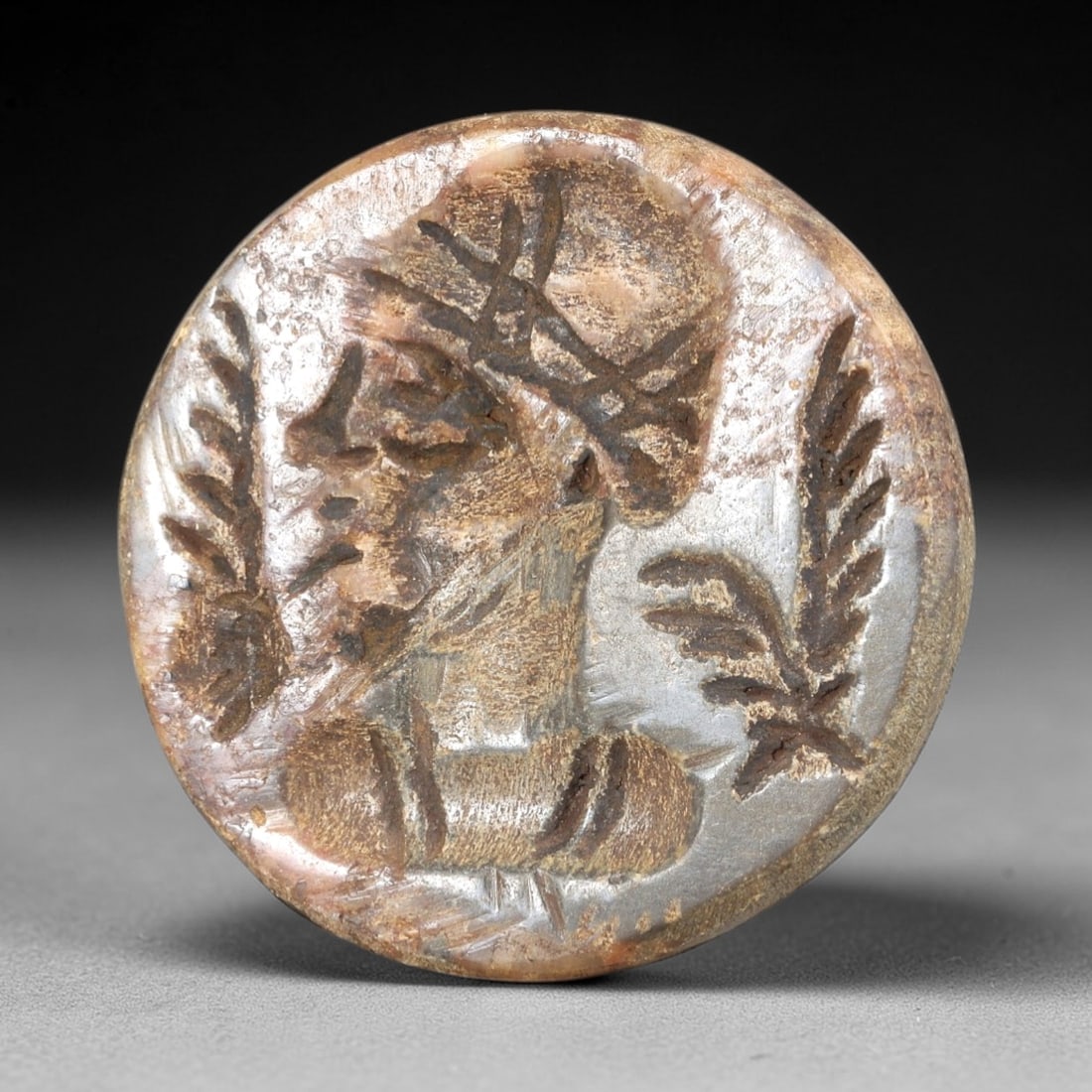 Ancient Stone Stamp Seal - Near Eastern Bust Figurine, 50AS: This ancient Near Eastern stamp seal is carved from stone, formed as a circular disc with a relief bust of a bearded male figure adorned with a spiked headdress, flanked by two stylized palm frond mot