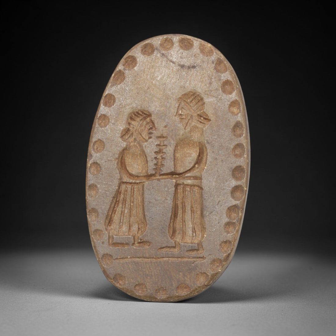 Ancient Stone Stamp Seal - Near Eastern Anthropomorphic Figurine, 68ZT: This ancient Near Eastern stamp seal is carved from stone, formed as an oval plaque with incised decoration depicting two confronting anthropomorphic figures in profile, jointly holding a stacked ritu
