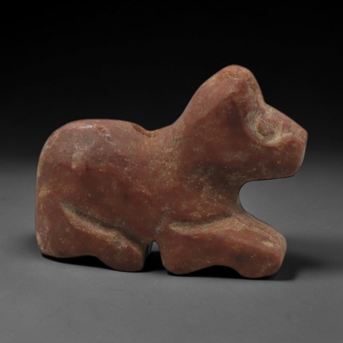 Ancient Stone Bovid - Near Eastern Amulet Pendant, 10XR: Carved from warm reddish-brown stone, this amulet depicts a stylized recumbent bovid with simplified facial features and a dorsal suspension hole. The abstract, geometric form is characteristic of Anc