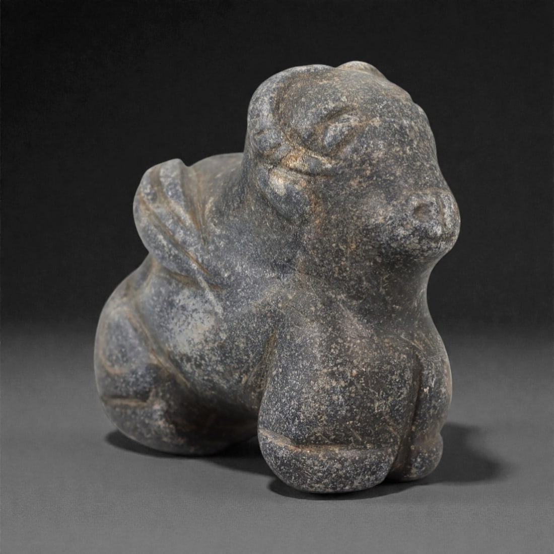 An Ancient Near Eastern Stone Zoomorphic Effigy, 3rd-1st Millennium BCE, 68FL: Carved from dark grey stone with light grey and brownish surface encrustation, the effigy depicts a stylized quadrupedal animal in a crouched pose. The head is rounded with recessed eye sockets, a pro