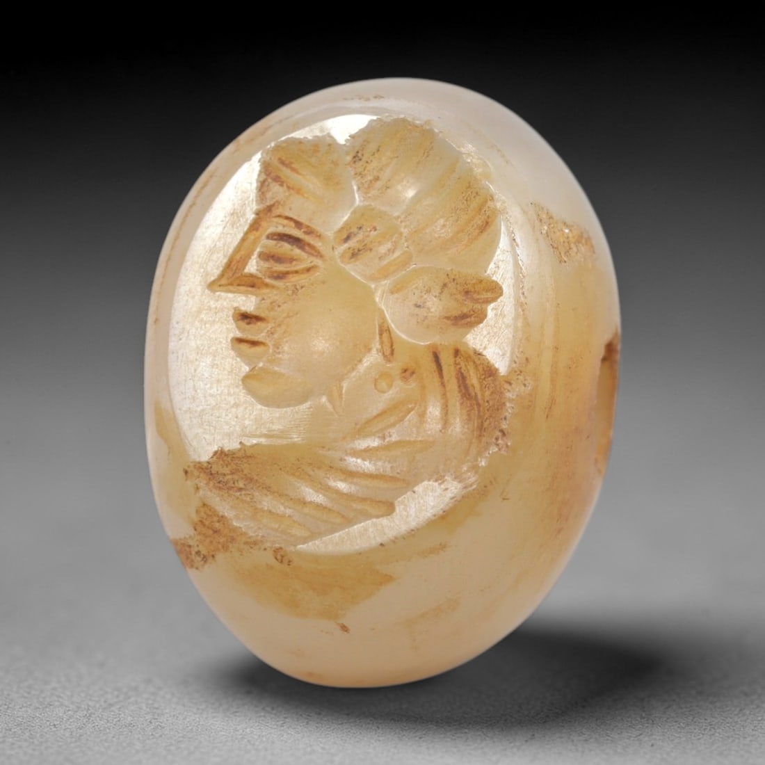 Ancient Chalcedony Stamp Seal - Near Eastern Female Bust, 66ZS: Crafted from translucent chalcedony, this oval stamp seal bears a deeply incised profile of a female bust, rendered with linear detailing to define her elaborate headdress, facial features, and draped