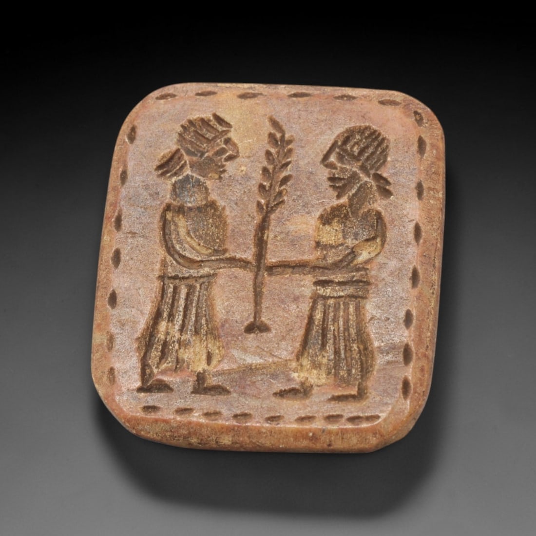 Ancient Steatite Stamp Seal - Near Eastern Anthropomorphic Figurine: This ancient Near Eastern stamp seal is carved from steatite, formed as a rounded rectangular plaque with incised decoration depicting two confronting anthropomorphic figures in profile, jointly holdi