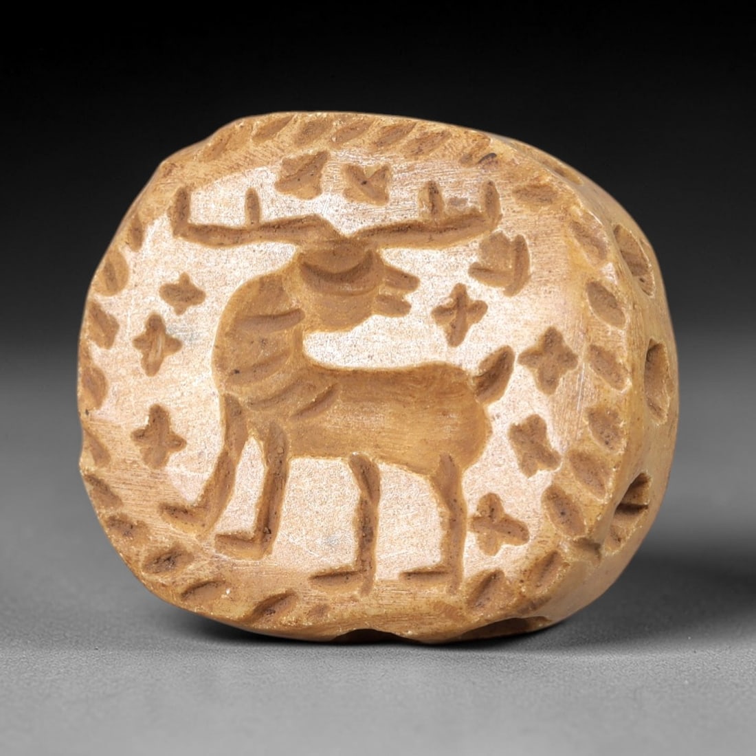 Ancient Steatite Stamp Seal - Near Eastern Stag Amulet: This ancient Near Eastern stamp seal is carved from steatite, formed as a rounded bead-like object with multiple suspension holes, featuring a relief depiction of a standing stag at its center, surrou
