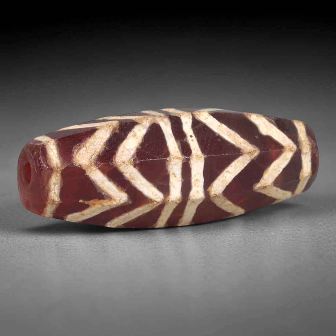 Ancient Carnelian Etched Bead - Himalayan Chevron Amulet, 99AR: This ancient elongated bead is crafted from polished carnelian, featuring a deep reddish-brown base with bold white etched chevron motifs across its multi-faceted surface. The bead retains a longitudi